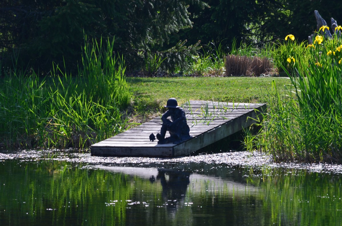 A classic sculpture of a boy fishing is situated at the end of a pond's dock.