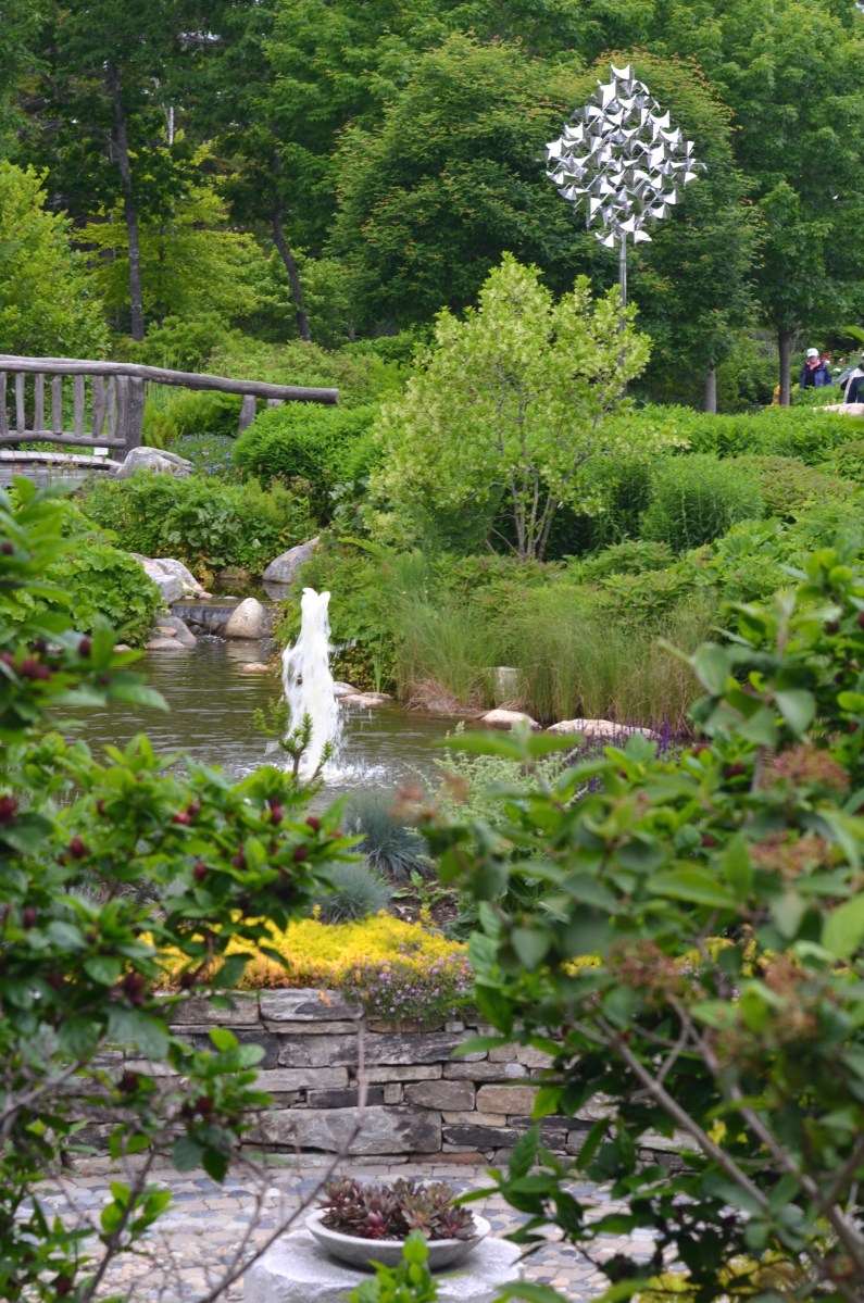 A pond, fountain and bridge with a kinetic sculpture in the background