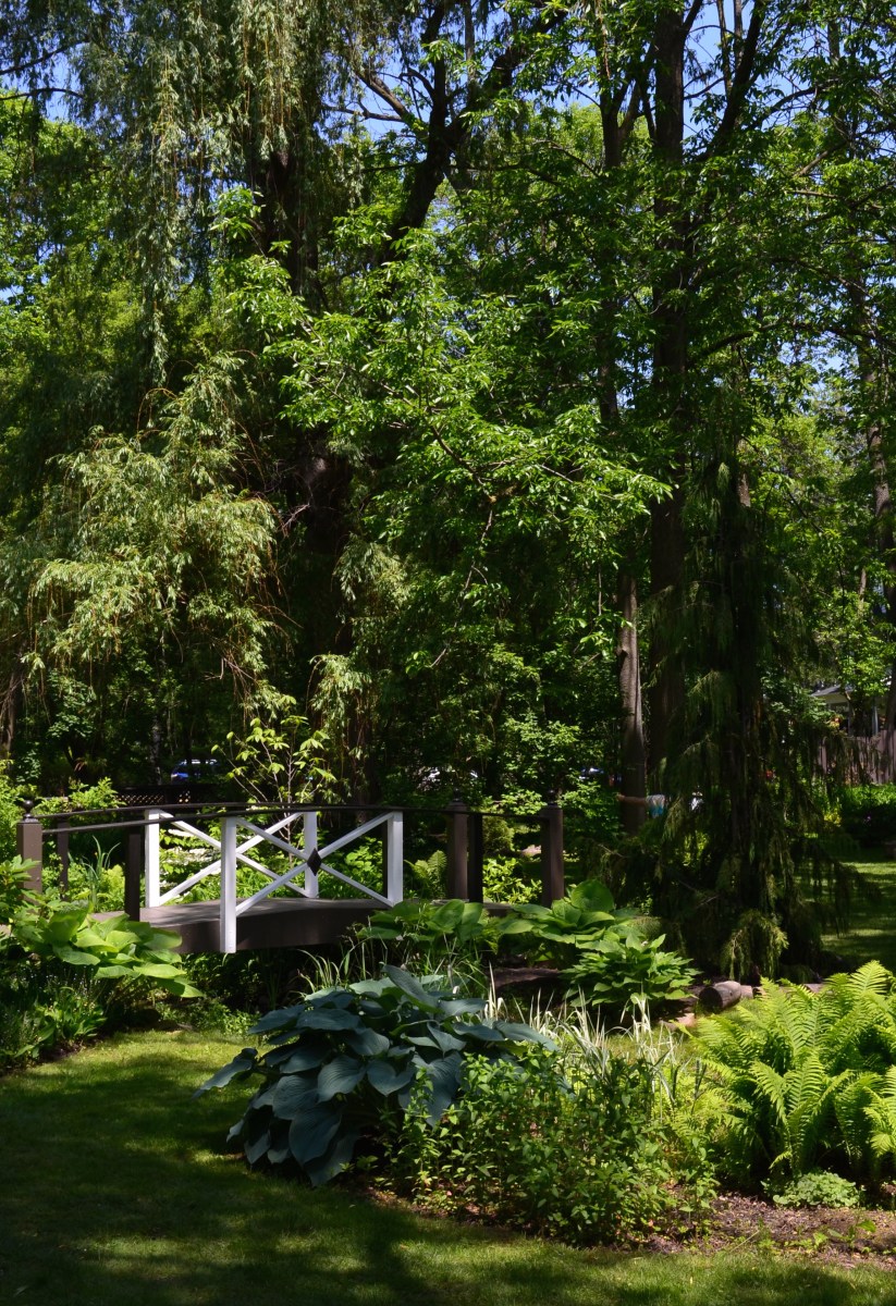 A white bridge spans a small creek in a garden with a woodland theme