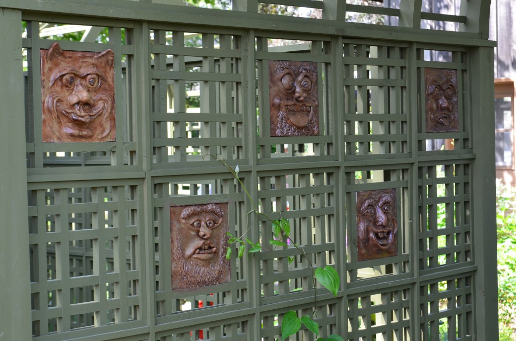 A collection of ceramic faces line the interior of a latticed archway in a garden.