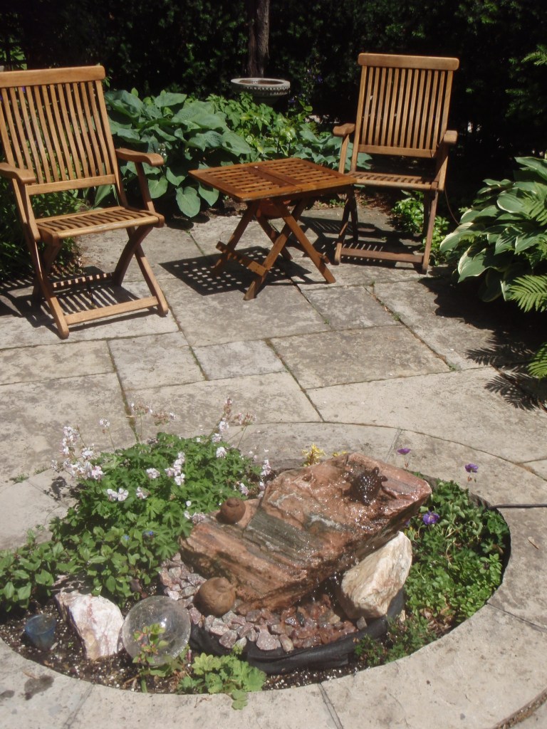 Two folding wooden chairs provide garden seating next to a small water feature in an urban garden.